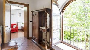 A room with a wooden ceiling, a red sofa, a large arched window with a view of greenery, and a wooden cabinet. - Ciliegio by Interhome (Castellina in Chianti)