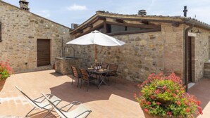 A stone building with a patio area featuring a table and chairs, an outdoor grill, and a potted plant with flowers. - Ciliegio by Interhome (Castellina in Chianti)