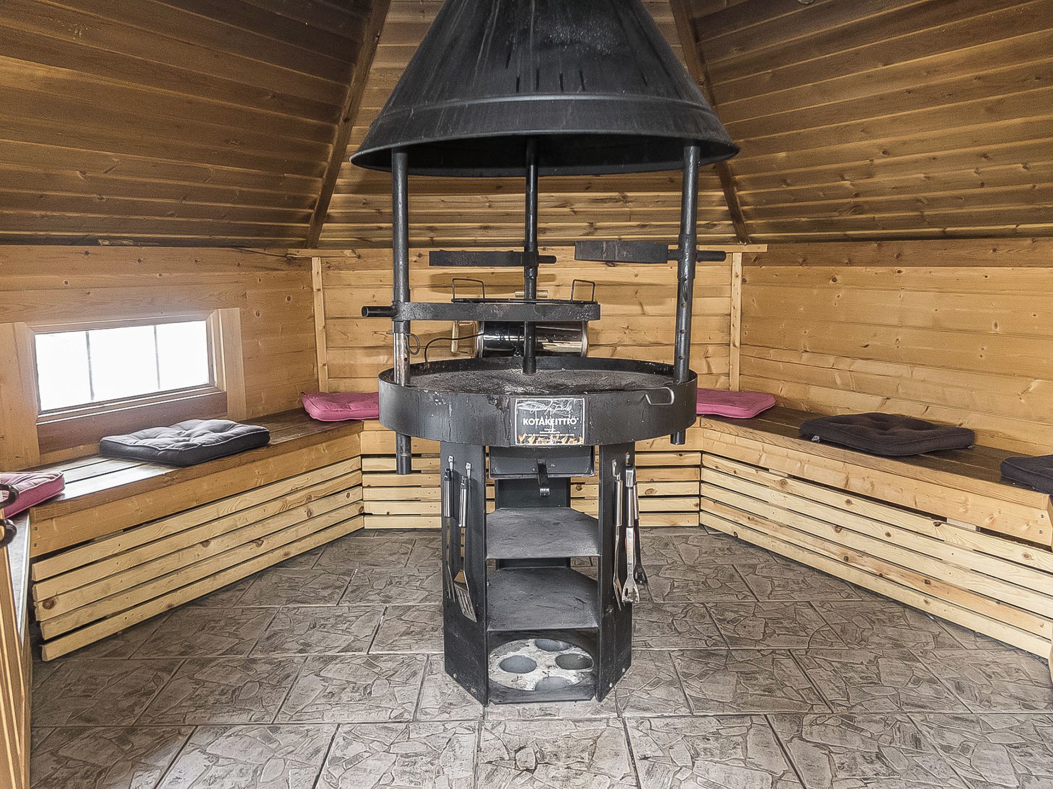 Un interior de fusta amb sòl de rajola de pedra, una estufa al centre i una finestra.