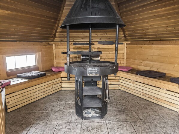 A wooden interior with a stone-tiled floor, a central stove, and a window.