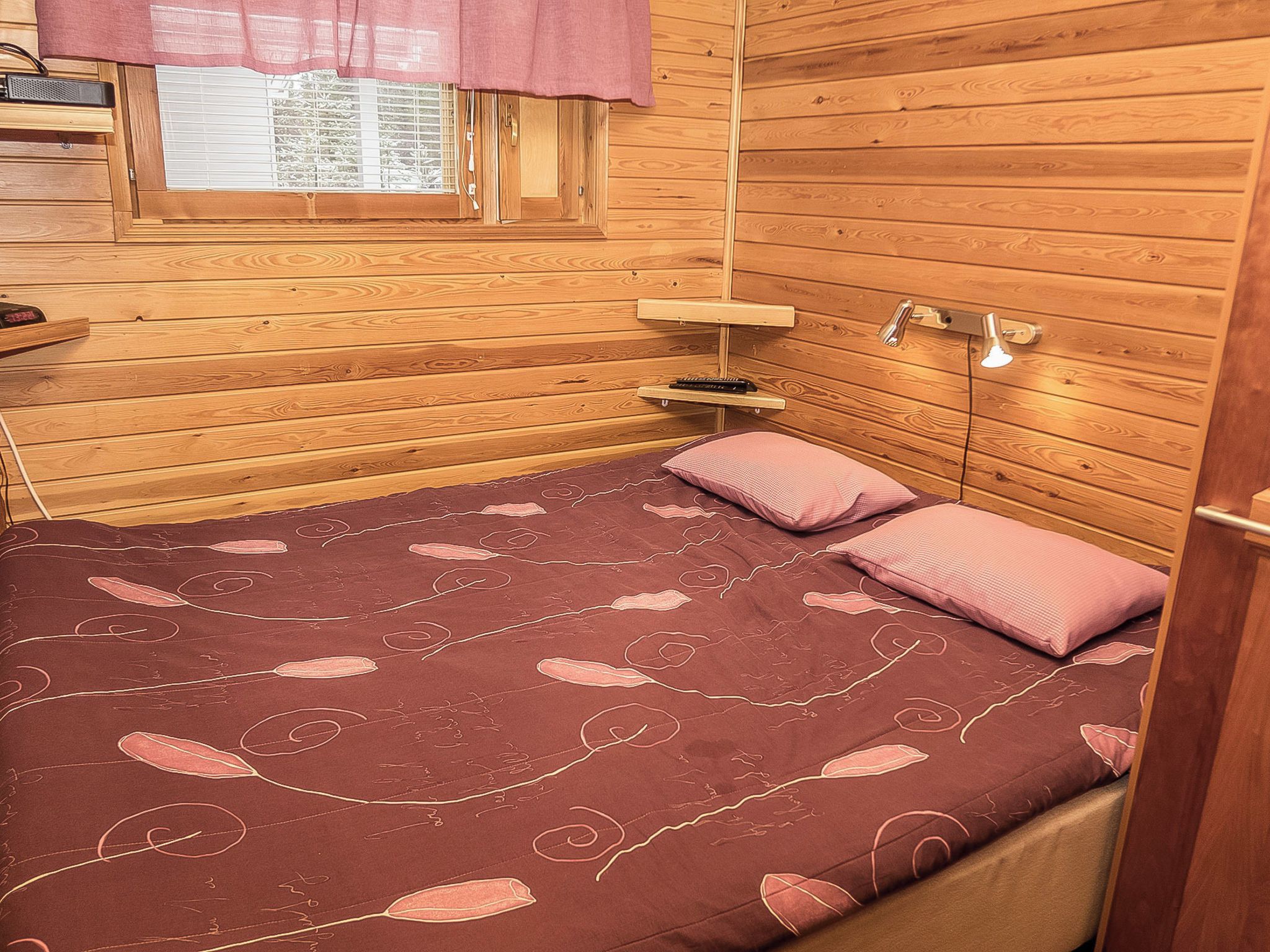 A wooden cabin bedroom with a bed, pillows, a nightstand, a window with blinds, and a wall-mounted lamp.