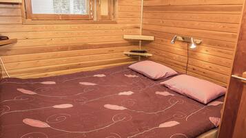 A wooden cabin bedroom with a bed, pillows, a nightstand, a window with blinds, and a wall-mounted lamp.