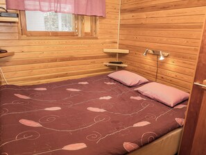 A wooden cabin bedroom with a bed, pillows, a nightstand, a window with blinds, and a wall-mounted lamp.