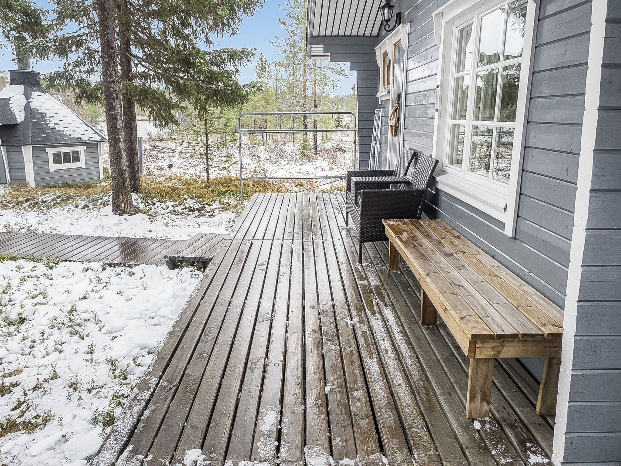 A wooden deck with a bench and a table, surrounded by snow-covered ground and trees.