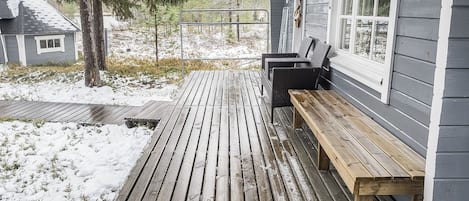 Een houten terras met een bankje en een tafel, omringd door sneeuw bedekte grond en bomen.