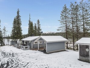 A cabin with a covered porch and a separate garage, surrounded by snow-covered ground and tall pine trees.