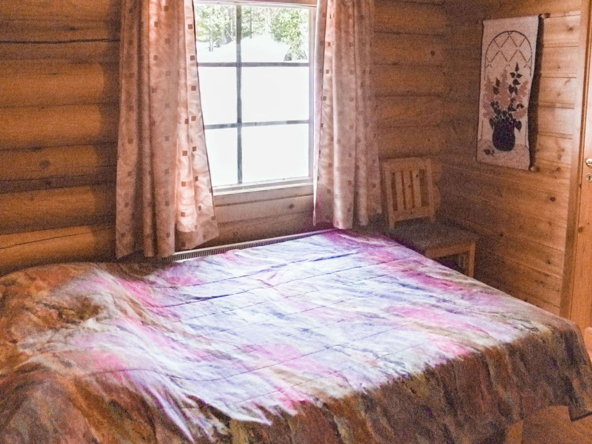 Une pièce de cabane en bois comprenant un lit, une fenêtre avec des rideaux, une chaise et un attrape-rêves fixé au mur.