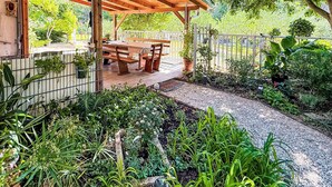 En overdækket terrasse med træbænke og et bord, omgivet af en have med forskellige planter og en grussti.