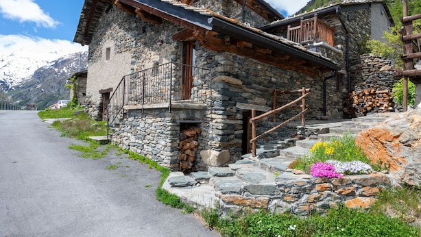 Ein Steinhaus mit einem Holzbalkon, einer Steintreppe und einem Berg im Hintergrund.