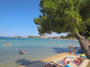 A beach with clear water, people swimming and sunbathing, and a tree providing shade.