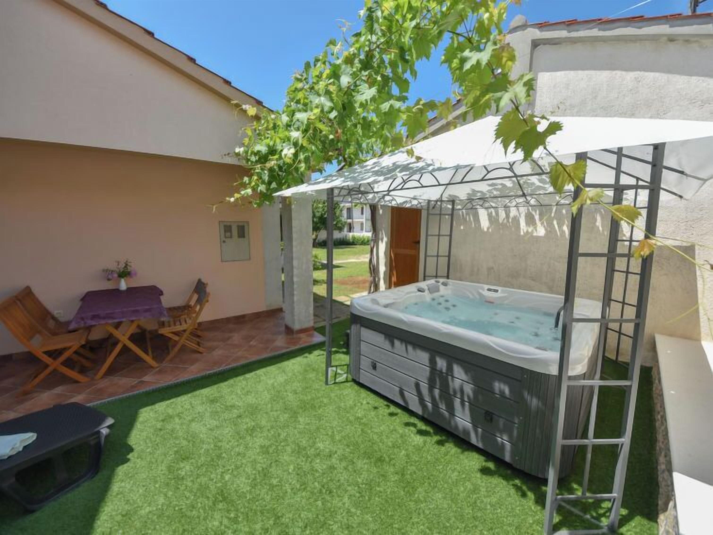 A patio with a hot tub, artificial grass, a table with chairs, and a pergola with vines.
