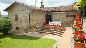 Une maison en pierre avec un toit en tuiles, un petit porche à l’avant avec des chaises en plastique blanches, et un jardin avec des fleurs rouges.