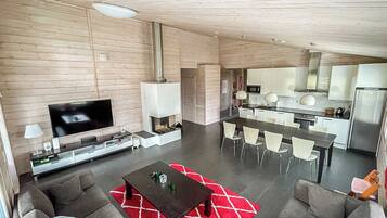 A modern living room with a flat-screen TV, a coffee table, and a dining area with chairs and a kitchen in the background.