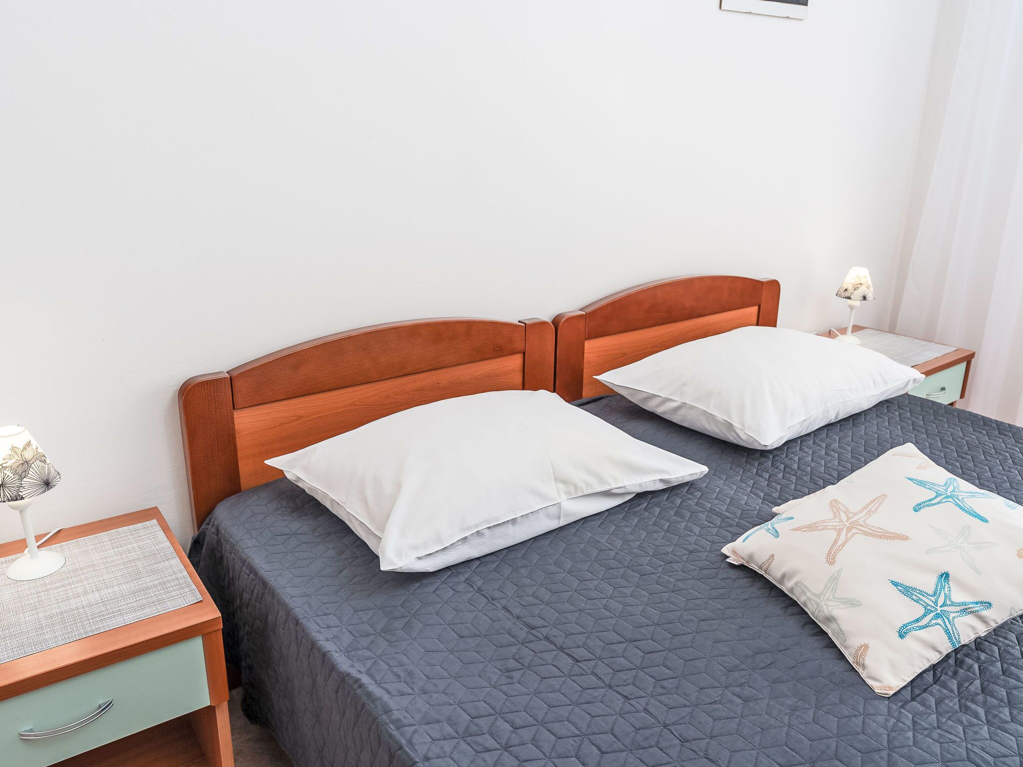 Ein ordentlich bezogenes Bett mit einer gesteppten, grauen Tagesdecke, zwei weißen Kissen und einem Kissen mit einem Seesternmuster. Ein hölzerner Kopfteil sowie ein Nachttisch mit Lampe und einer kleinen Schublade sind ebenfalls zu sehen.