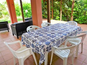 Une terrasse extérieure avec une table recouverte d’une nappe à motifs floraux bleus et blancs, entourée de chaises en plastique blanches.