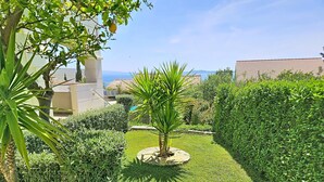 A well-maintained garden with a palm tree, neatly trimmed hedges, and a clear view of the sea in the background.