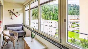 En balkon med en grå sofa, en fletstol og et lille bord med en potteplante. Store vinduer giver udsigt over et skovområde og bygninger.