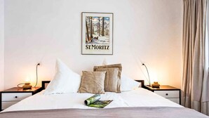En pænt redt seng med et indrammet billede af St. Moritz over, to sengeborde med lamper, og et vindue med gardiner.