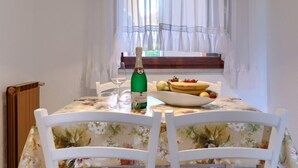 Une table à manger recouverte d’une nappe à motifs floraux, avec des chaises blanches, une bouteille de champagne, un verre à vin et un saladier rempli de fruits.