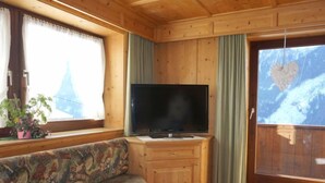 Een gezellig chaletinterieur met een houten plafond, een televisie op een houten kast, een beklede bank en een raam met gordijnen.