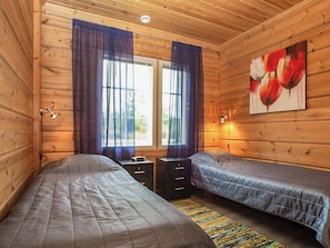 Une chambre dans un chalet en bois, avec deux lits, une fenêtre avec des rideaux, un tableau et une table de chevet.