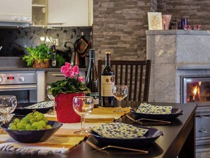 Une table à manger dressée avec des verres à vin, une bouteille de vin, un bol de raisins et un pot rouge contenant des fleurs.