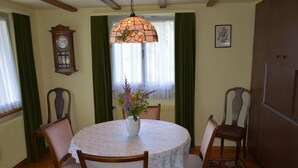 Un comedor con una mesa redonda, mantel floral, cuatro sillas de madera, un jarrón con flores, una lámpara de cristal y un reloj de pared, además de un cuadro enmarcado.