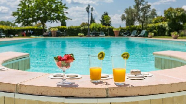 Escena junto a la piscina, con vista clara al agua, un vaso de zumo de naranja y un bol de fresas.