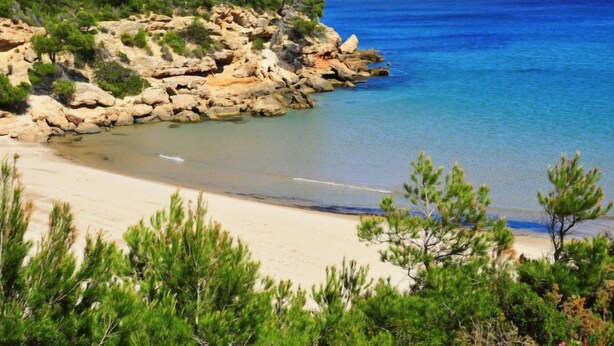 A coastal landscape with a rocky shoreline, clear blue sea, and sandy beach.