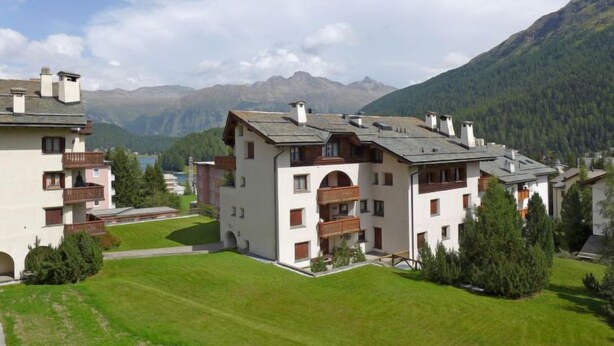 A multi-story building with balconies, surrounded by lush greenery and mountains in the background.