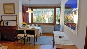 A room with a dining table set for two, a wooden cabinet, a large window with a view of buildings and trees, and a painting on the wall.