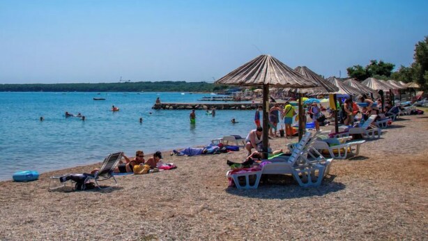A beach with sun loungers and umbrellas, a pier in the background, and people swimming and relaxing. - Graziani by Interhome (Krk/Kornic)