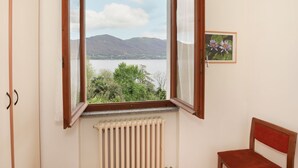 A room with a window overlooking a lake and mountains, a radiator below the window, and a chair to the right. - Rossella by Interhome (Castelveccana)