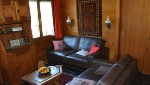 Een gezellig interieur van een chalet met een leren bank, een houten eettafel met fruit, en een groot vloerkleed.