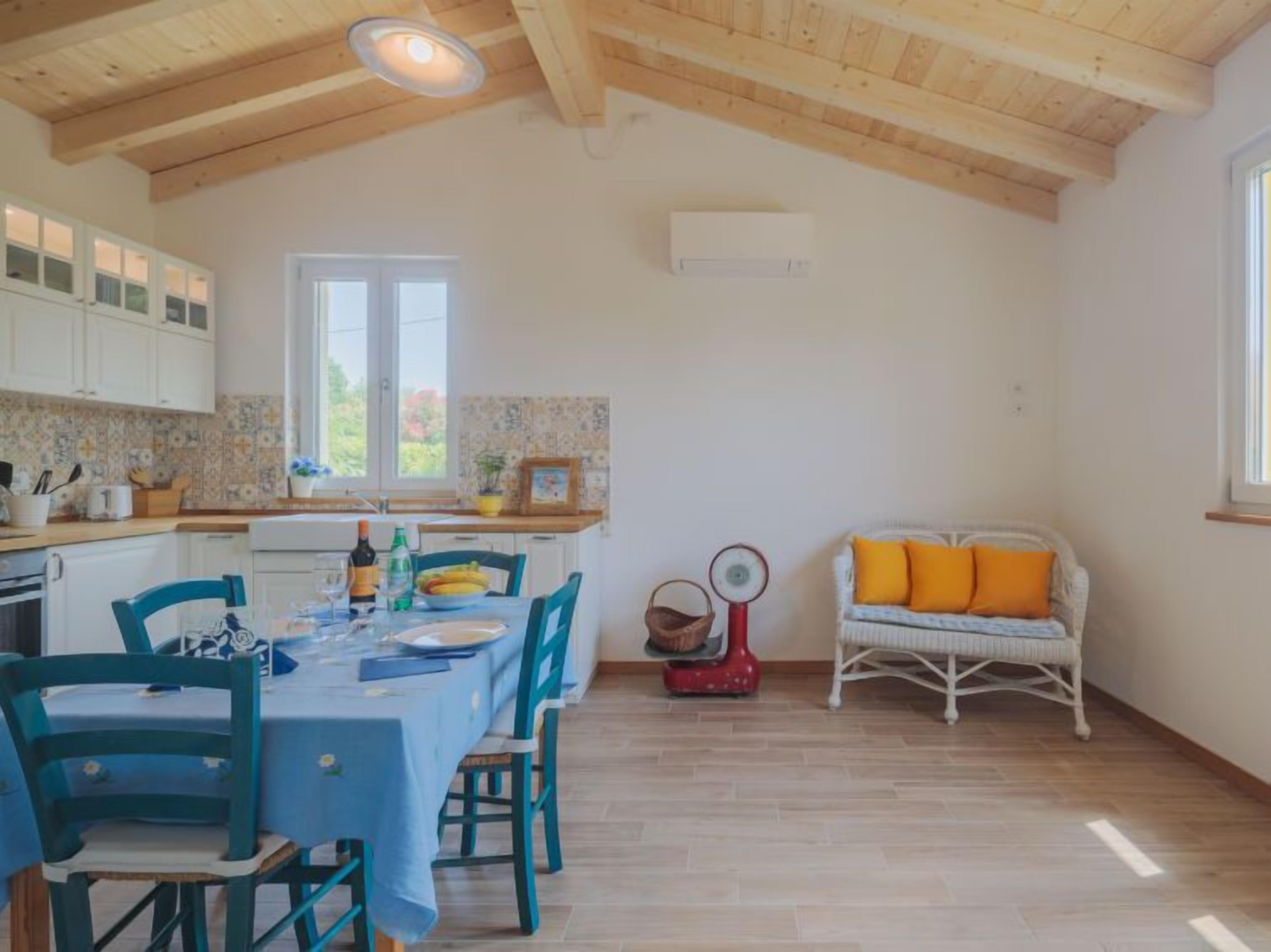 Un'ampia cucina con travi in legno, un'area pranzo con tavolo e sedie, un divano in vimini con cuscini arancioni e una finestra che offre luce naturale.