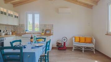 Un'ampia cucina con travi in legno, un'area pranzo con tavolo e sedie, un divano in vimini con cuscini arancioni e una finestra che offre luce naturale.