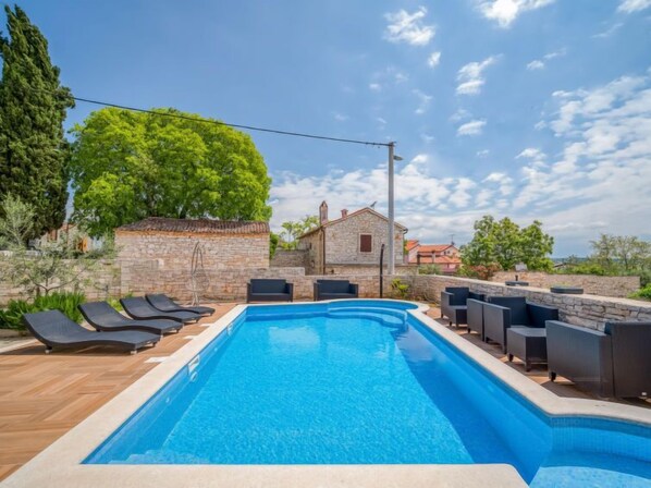 Piscina retangular com borda de pedra, cercada por espreguiçadeiras e um muro de pedra.