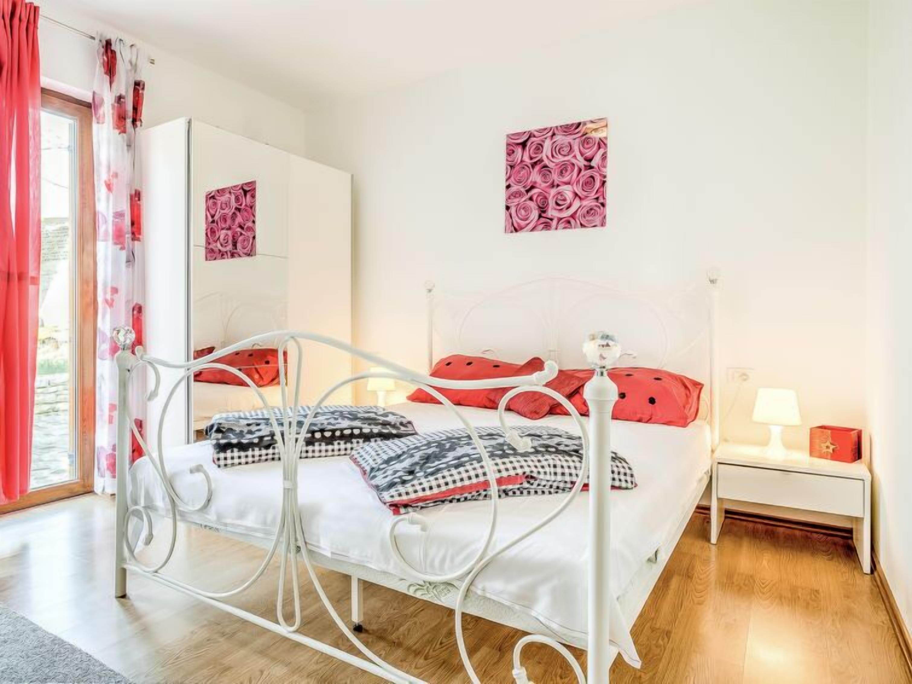 Une chambre à coucher avec un lit en métal, une literie blanche, des coussins rouges, une table de chevet avec une lampe et un mur orné d’une œuvre d’art florale.