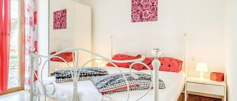 Une chambre à coucher avec un lit en métal, une literie blanche, des coussins rouges, une table de chevet avec une lampe et un mur orné d’une œuvre d’art florale.