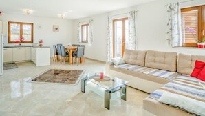 A spacious living area with a sofa, a glass coffee table, and a dining table with chairs. There is a kitchen area with a refrigerator and a window.