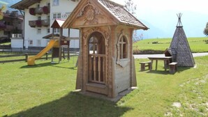 A small wooden structure with a sloped roof and decorative elements, situated in a grassy area with a playground and a building in the background. - Schwendau by Interhome (Mayrhofen)
