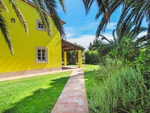 A yellow building with a brick pathway leading to a covered area, surrounded by greenery and palm trees.