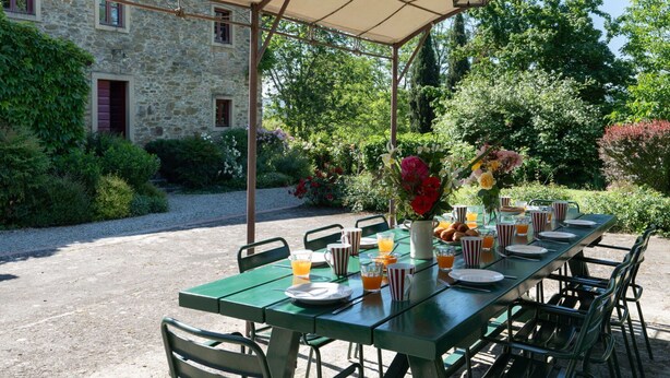 An outdoor dining area with a table set for breakfast, surrounded by greenery and a stone building. - La Forra by Interhome (Vicchio)