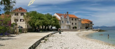 A coastal building with a stone facade and red-tiled roofs, situated on a pebbled beach with clear water and distant mountains.