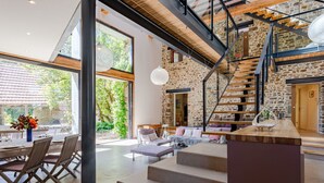 Un intérieur spacieux avec un escalier menant à une mezzanine, un coin repas offrant une vue agréable et un espace salon confortable agrémenté de coussins.