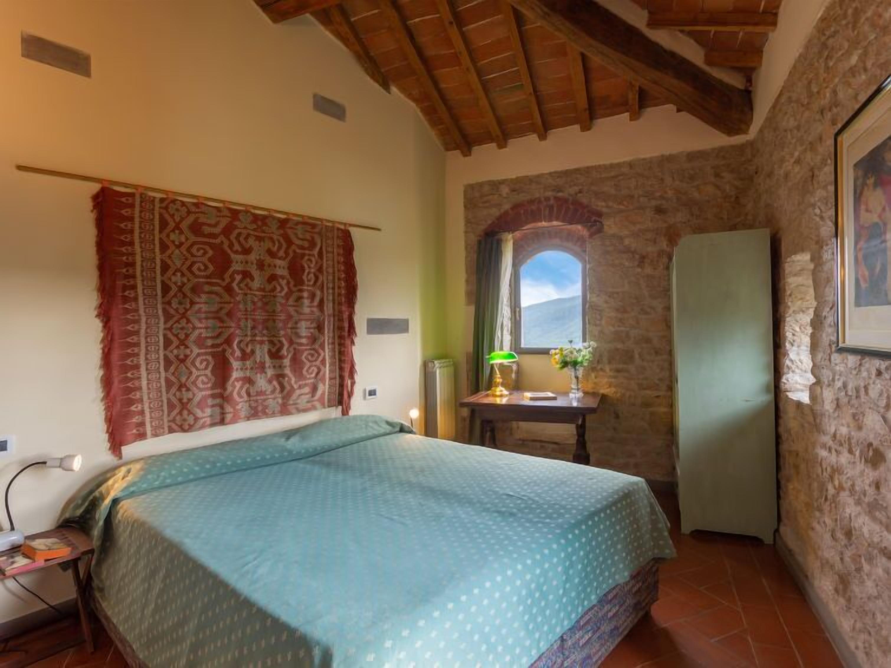 A bedroom with a bed, a desk with a lamp, a window, and a refrigerator.