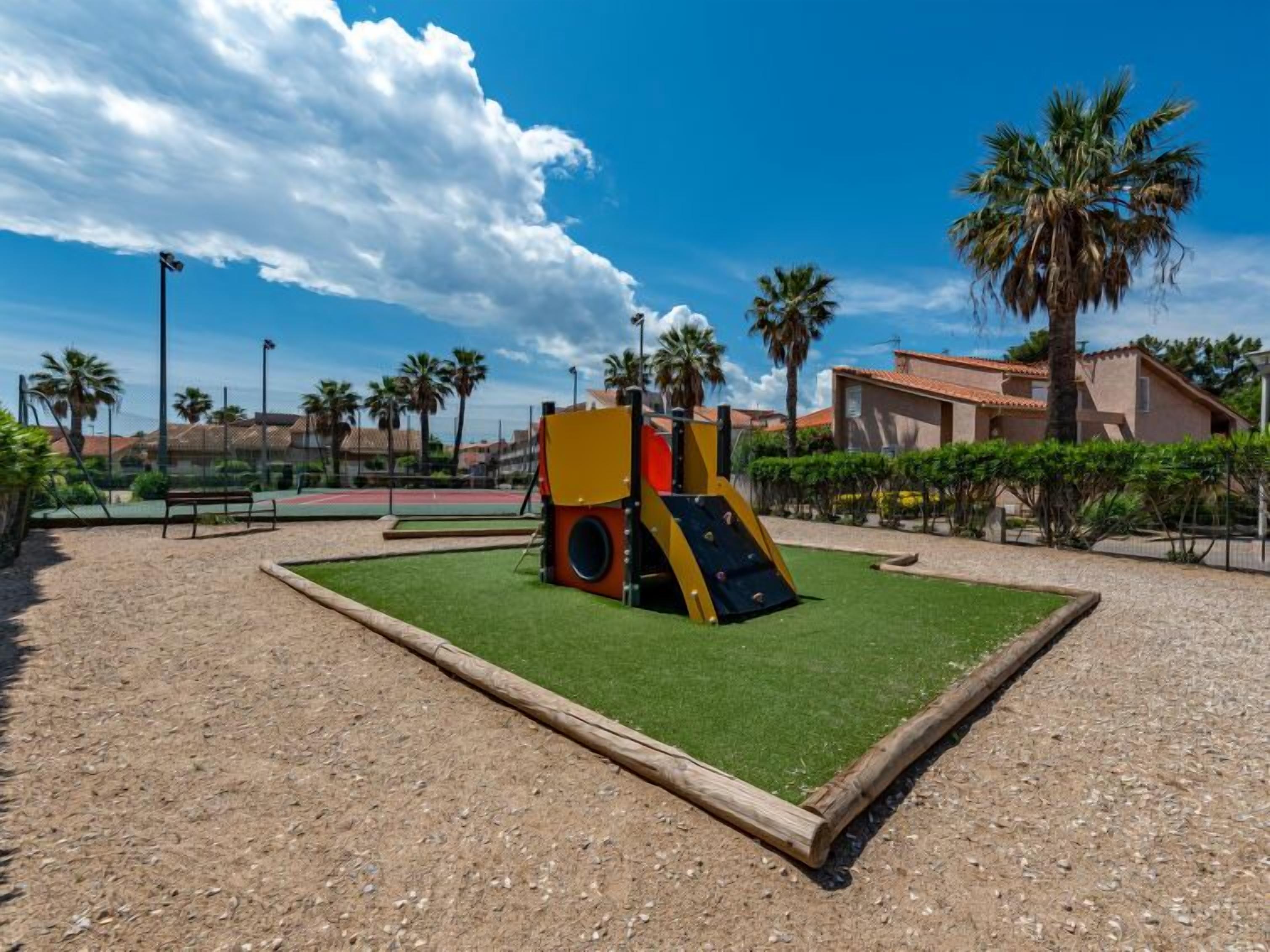 A playground with a slide and climbing wall, surrounded by palm trees and a tennis court.