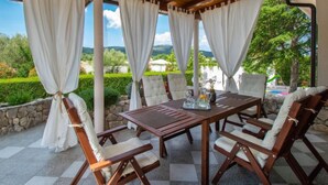 Un patio al aire libre con una mesa y sillas de madera, rodeado de cortinas blancas, con vistas panorámicas a zonas verdes y colinas.