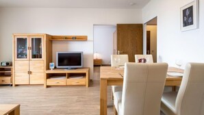 Un salon moderne avec un meuble en bois, un téléviseur à écran plat, une table à manger avec des chaises blanches et un tableau encadré au mur.