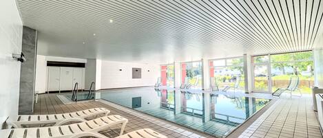 Een modern binnenzwembad met glazen wanden, witte lounge stoelen en een betegelde vloer.
